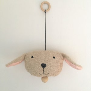 Può includere: Un mobile a forma di testa di cane in peluche beige e rosa con occhi e naso ricamati neri. Il mobile è appeso a un anello di legno e una corda nera.
