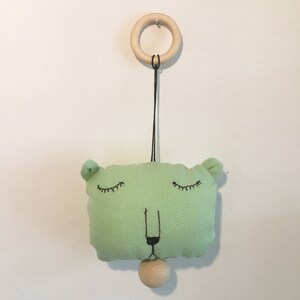 May include: A green fabric toy with a face, closed eyes, and a small wooden ball hanging from it. The toy is attached to a wooden ring by a black string.