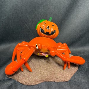 May include: A Halloween-themed figurine featuring a bright orange crab with outstretched claws, supporting a carved pumpkin on its back. The pumpkin has a smiling face and green stem. The crab rests on a brown, textured base.