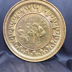 May include: A round, antique brass plate with an embossed scene of people gathered around a table. The plate has a decorative border with a repeating leaf design. The diameter of the plate is approximately 12 inches.