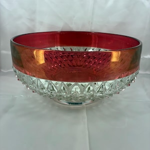 May include: A round glass bowl featuring a deep red upper band and a clear, faceted lower section with a diamond-cut pattern. The interior of the bowl reflects the red hue, while the exterior base displays a textured, geometric design.