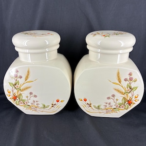 May include: Two off-white ceramic jars with lids. Each jar features a floral design with wheat, berries, and leaves in shades of brown, green, and red. The jars have a rounded shape and are set against a dark background.