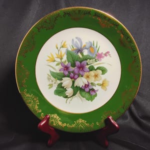 May include: A decorative plate with a green border and a gold filigree design. The center features a floral bouquet with purple, yellow, white, and blue flowers. The plate is displayed on a red stand.