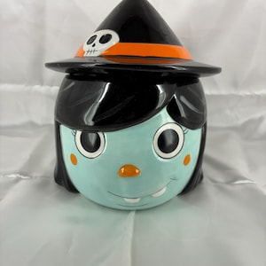 May include: A ceramic cookie jar shaped like a witch's head with light blue skin, black hair, and a black pointed hat. The hat features an orange band and a small white skull accent. The face has large black eyes, orange cheeks, and a single tooth.