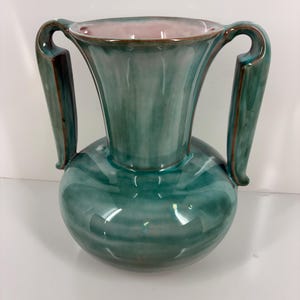 May include: A teal ceramic vase with two curved handles. The vase has a bulbous base and a flared top with a pink interior. The handles are a darker teal with brown accents. The vase is a decorative item.