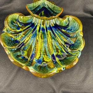 May include: A ceramic seashell-shaped dish with a glossy finish. The dish features a blend of green, blue, and yellow hues, with a brown rim. The design includes detailed ridges and a scalloped edge, creating a decorative and functional piece.