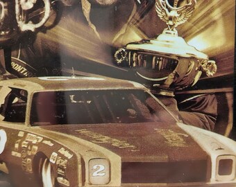 May include: A sepia-toned image featuring Dale Earnhardt, holding a trophy, with a race car in the foreground. The car is number 2. Text at the bottom reads "Dale Earnhardt. Continue The Legend. First Championship - 1980."
