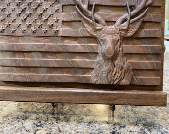May include: A wooden wall art piece featuring a carved American flag with a deer head in the center. The deer has large antlers and a detailed face. The piece is mounted on a wooden frame.