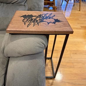 May include: A wooden side table with a black metal frame. The tabletop is rectangular, made of light brown wood, and features a black decorative design. The table is designed to slide over the arm of a sofa or chair.