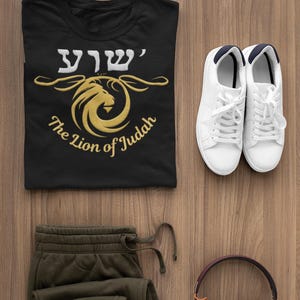 May include: Black t-shirt with a gold lion graphic and the text "The Lion of Judah". The image also shows olive green sweatpants, white sneakers, and maroon headphones, all arranged on a wooden surface.