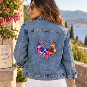 May include: A blue denim jacket featuring a vibrant graphic on the back. The design displays two large butterflies, one in shades of purple and blue and the other in orange and red, surrounded by small colorful flowers and foliage accents.