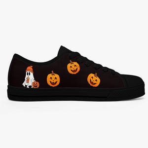 May include: Black canvas sneakers with a Halloween theme. The design features a cartoon ghost wearing an orange beanie and holding a drink, along with several smiling jack-o'-lanterns. The shoes have black laces and soles.