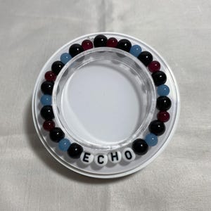 May include: A beaded bracelet with black, red, and light blue beads. The bracelet is on a white circular base with the word "ECHO" spelled out in white letter beads.