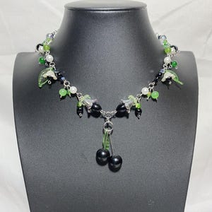 May include: A silver chain necklace with black and green beads, white pearls, and silver bells. The necklace has a dangling charm of two black cherries with green stems.