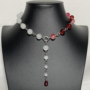 Puede incluir: Un collar de cuentas con un diseño degradado. El collar hace la transición de cuentas blancas a rosas y rojas. Una hebra vertical de cuentas blancas cuelga del centro, terminando con una cuenta roja en forma de lágrima. El collar se exhibe en un maniquí negro.
