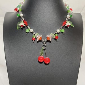 May include: A delicate necklace featuring a silver chain adorned with red, green, and white beads. The necklace includes clear flower and leaf charms, and a pair of red cherry pendants. The necklace is displayed on a black mannequin.