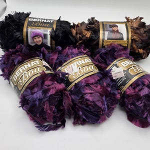 May include: Close-up of several skeins of Bernat Boa yarn in various colors. The yarn is a fluffy, textured material, with colors including black, brown, and shades of purple. Each skein has a label with the brand name and a picture of a person.