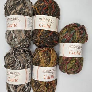 May include: Six skeins of Moda Dea Cache yarn in various colors. The yarn is a mix of brown, gold, and green with metallic threads. Each skein has a white label with the brand name and yarn name. Ideal for knitting and crochet projects.