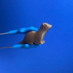 May include: A brown, 3D-printed otter figurine held by blue tools against a blue backdrop. The otter is in a standing pose, with its head and body slightly angled. The tools appear to be holding the otter securely.