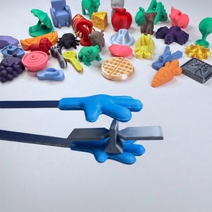 以下が含まれることがあります： 青い手形のピンセットが小さな灰色のT字型のプラスチック片を掴んでおり、背景にはワッフル、果物、動物、家庭用品など、さまざまなカラフルな3Dプリントされたミニチュアオブジェクトが散らばっています。