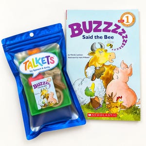 May include: A blue plastic pouch labelled Talkets by Speech & Smile containing small colourful items and a miniature book cover, alongside a Scholastic Level 1 reader book titled BUZZZZ Said the Bee featuring illustrations of a cow, pig, duck, and chicken.