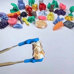 May include: A collection of small, multi-colored 3D printed plastic figurines including animals, food items, and objects. A pair of blue-tipped metal tweezers holds a single, shiny, cream-colored figurine of a person wearing a crown.