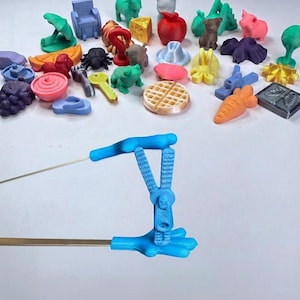 Puede incluir: Un tirador de cremallera de plástico azul con forma de mano que sostiene un cursor, unido a un alambre de metal. Al fondo, una colección de pequeños objetos de plástico impresos en 3D, incluyendo comida, animales y diversas formas.