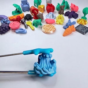 May include: A collection of small, colorful 3D-printed plastic figurines in various shapes including animals, food items, and objects, alongside a pair of blue plastic-tipped metal tongs holding a light blue 3D-printed wave-shaped figurine.