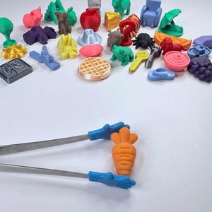 以下が含まれることがあります： 動物、食べ物、その他の様々な形をしたカラフルな3Dプリント製の小さなプラスチック製フィギュアのコレクションと、青い手形のシリコン製先端が付いた金属製のトングで、小さなオレンジ色のニンジン型のフィギュアを挟んでいます。