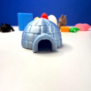 May include: A light blue 3D-printed igloo with a rounded entrance. The igloo is the focal point, with other colorful 3D-printed shapes in the background, including a blue block, a red cone, and a brown animal figure. The objects are on a white surface against a blue backdrop.