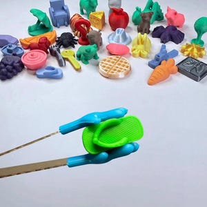 May include: A collection of small, multi-colored 3D printed plastic figurines and objects, including food items, animals, and household shapes. A pair of blue-tipped metal tweezers holds a single bright green plastic flip-flop sandal.