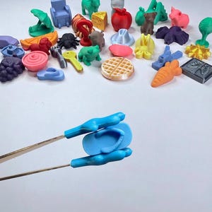 May include: A collection of small, multi-colored 3D printed plastic miniatures including animals, food items, and household objects. A pair of metal tweezers holds a single light blue flip-flop miniature in the foreground of the image.
