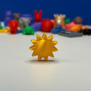 May include: A golden yellow, sun-shaped 3D-printed object. The sun has pointed rays and is the focal point of the image. Other colorful 3D-printed toys are in the background, out of focus.