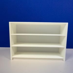 May include: A small, white, three-tiered shelf unit. The shelves are evenly spaced and the unit has a rectangular shape. The background is a solid blue color. The shelf is likely for displaying small items.