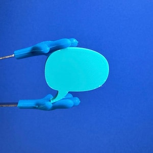 May include: A turquoise speech bubble is held by two blue plastic hands against a solid blue background. The hands are attached to thin metal rods. The speech bubble has a textured surface.