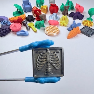 Puede incluir: Una colección de pequeñas figuras de plástico impresas en 3D, multicolores, que incluyen animales, alimentos y objetos. Unas pinzas de plástico azul sostienen una placa cuadrada de color gris oscuro con un diseño de caja torácica en relieve.