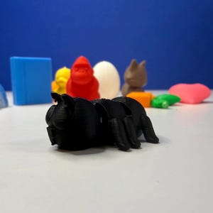 Puede incluir: Una araña de juguete negra, impresa en 3D, está en primer plano, con otros juguetes coloridos impresos en 3D en el fondo. Los otros juguetes incluyen un cubo azul, un pato amarillo, un gorila rojo, un huevo blanco, un conejo marrón, un bloque naranja, una tortuga verde y un corazón rosa.