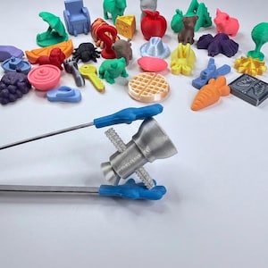 Puede incluir: Unas pinzas de metal con puntas de silicona azul en forma de mano sostienen un pequeño objeto plateado impreso en 3D. Al fondo, hay una colección de varias figuras pequeñas y coloridas de plástico impresas en 3D, incluyendo un gofre, frutas, animales y formas.