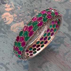 May include: A silver-toned bangle bracelet featuring a dense, alternating pattern of oval-shaped, vibrant pink and deep green gemstones. The metal frame has a textured, beaded edge, and the interior displays a repeating pattern of circular cutouts.
