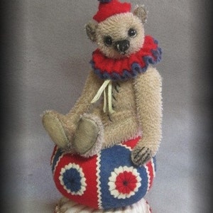 Puede incluir: Un oso de peluche beige con un gorro de buf&oacute;n rojo y azul y un cuello con volantes rojo y azul se sienta sobre una bola roja, blanca y azul. La bola est&aacute; sobre una base blanca con un borde festoneado rojo.