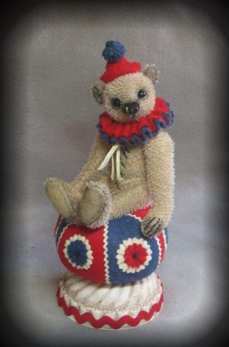 Puede incluir: Un oso de peluche beige con un gorro de buf&oacute;n rojo y azul y un cuello con volantes rojo y azul se sienta sobre una bola roja, blanca y azul. La bola est&aacute; sobre una base blanca con un borde festoneado rojo.
