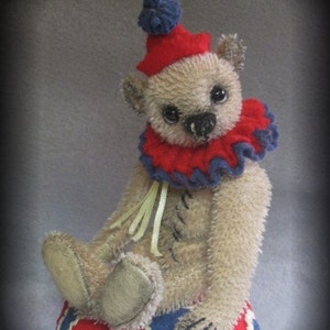Puede incluir: Un oso de peluche beige con un gorro de buf&oacute;n rojo y azul y un cuello de volantes rojo y azul. El oso est&aacute; sentado en una pelota con estampado rojo, blanco y azul.