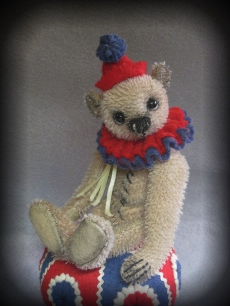 Puede incluir: Un oso de peluche beige con un gorro de buf&oacute;n rojo y azul y un cuello de volantes rojo y azul. El oso est&aacute; sentado en una pelota con estampado rojo, blanco y azul.