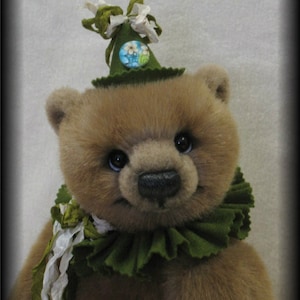 Puede incluir: Un oso de peluche marrón que lleva un sombrero de fieltro verde con un botón floral y un cuello de volantes de fieltro verde. El oso tiene una cinta blanca atada alrededor del cuello.