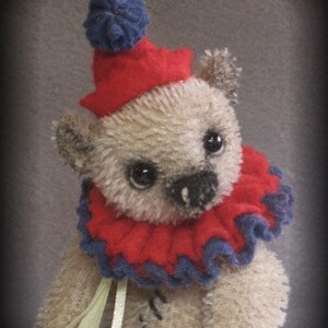 Puede incluir: Un oso de peluche beige que lleva un gorro de buf&oacute;n rojo y azul con un pomp&oacute;n azul en la parte superior. El oso tiene un cuello con volantes rojo y azul y sostiene una cinta amarilla.