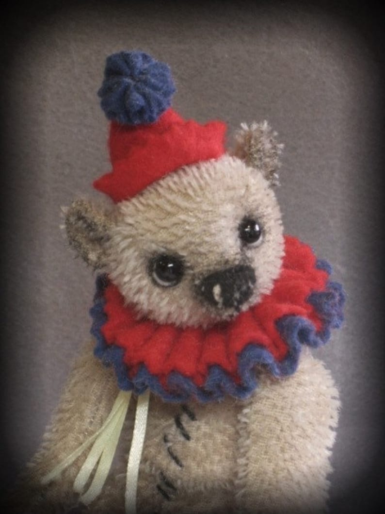Puede incluir: Un oso de peluche beige que lleva un gorro de buf&oacute;n rojo y azul con un pomp&oacute;n azul en la parte superior. El oso tiene un cuello con volantes rojo y azul y sostiene una cinta amarilla.
