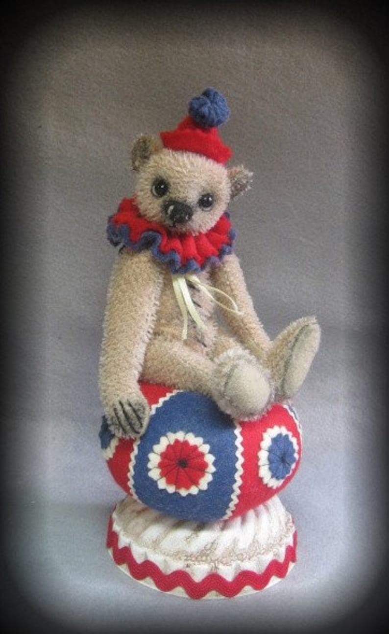 Puede incluir: Un oso de peluche beige con un traje de payaso rojo y azul se sienta sobre una bola roja, blanca y azul. El oso lleva un sombrero azul y blanco y una cinta blanca alrededor del cuello.