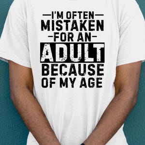 Puede incluir: Camiseta blanca con el texto "-I'M OFTEN- MISTAKEN -FOR AN- ADULT BECAUSE OF MY AGE" en negro. La camiseta la lleva una persona de piel oscura, y se ve un reloj negro en su mu&ntilde;eca.