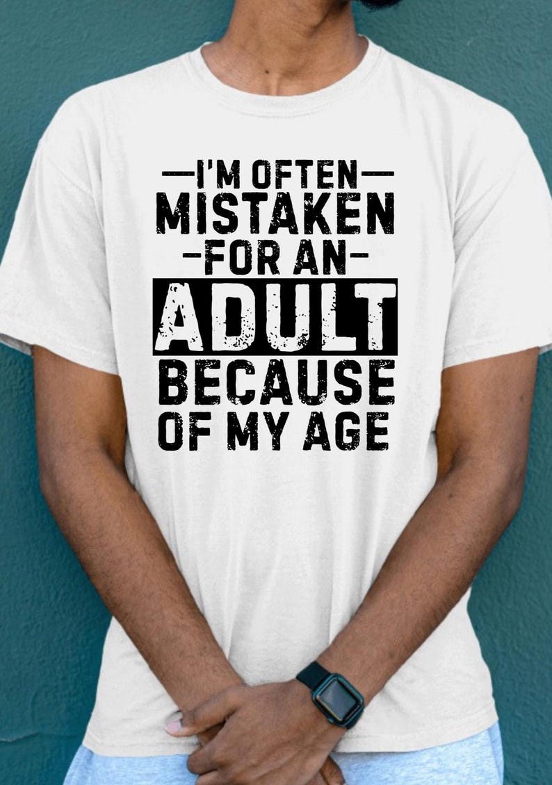 Puede incluir: Camiseta blanca con el texto "-I'M OFTEN- MISTAKEN -FOR AN- ADULT BECAUSE OF MY AGE" en negro. La camiseta la lleva una persona de piel oscura, y se ve un reloj negro en su mu&ntilde;eca.