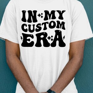 K&ouml;nnte beinhalten: Wei&szlig;es T-Shirt mit schwarzem Text, der "IN MY CUSTOM ERA" lautet, mit Sternen und Blitzen um den Text herum.